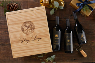 A wooden box with "Stags' Leap" Winery branding sits beside three wine bottles, a corkscrew, a pine cone, and a wrapped gift on a rustic wooden surface.